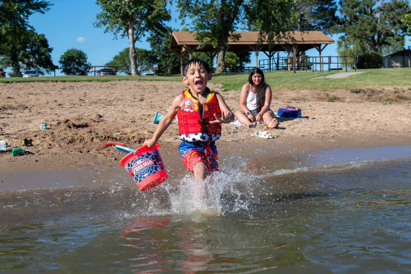A small boy wearing a lifejacket runs into the lake carrying a sand bucket