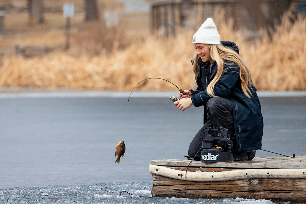 A smiling woman reels in a bluegill while ice-fishing off a wood dock during winter.