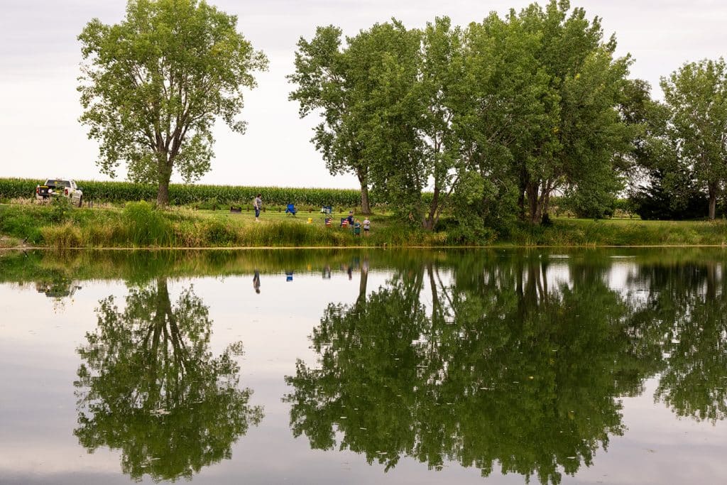 A family fishes at a pit lake lined with trees reflecting on calm water during summertime.