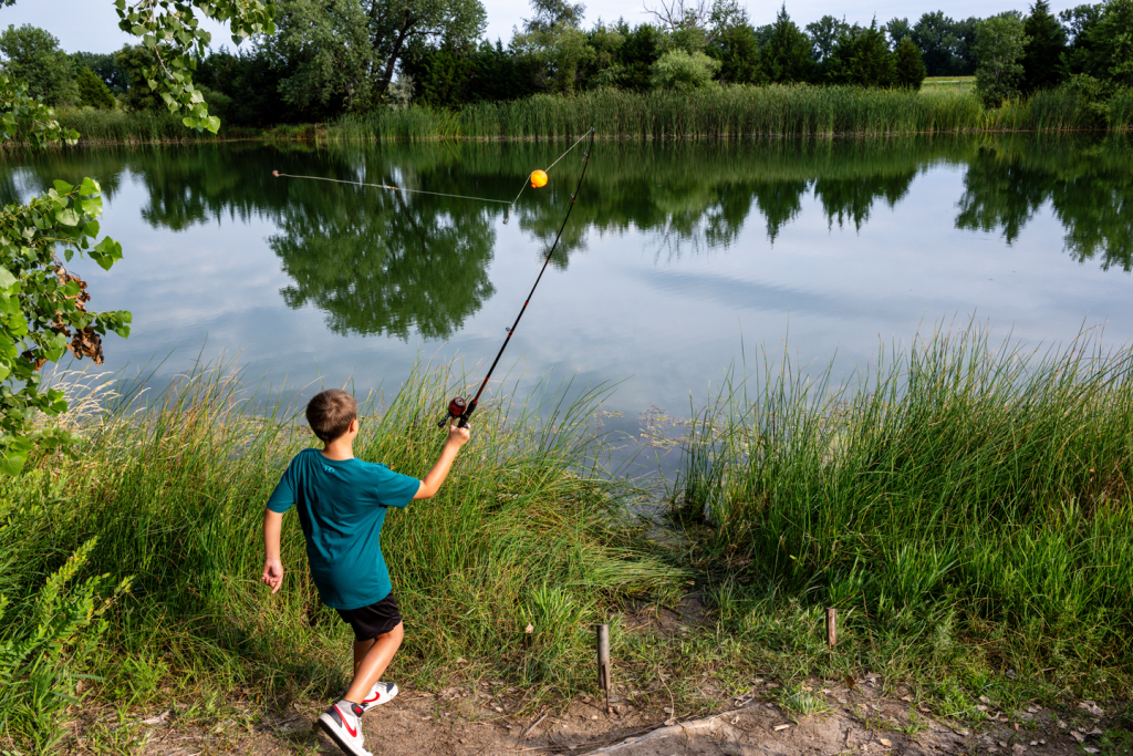 A boy fishes on the bank of a lake lined with trees during summer.