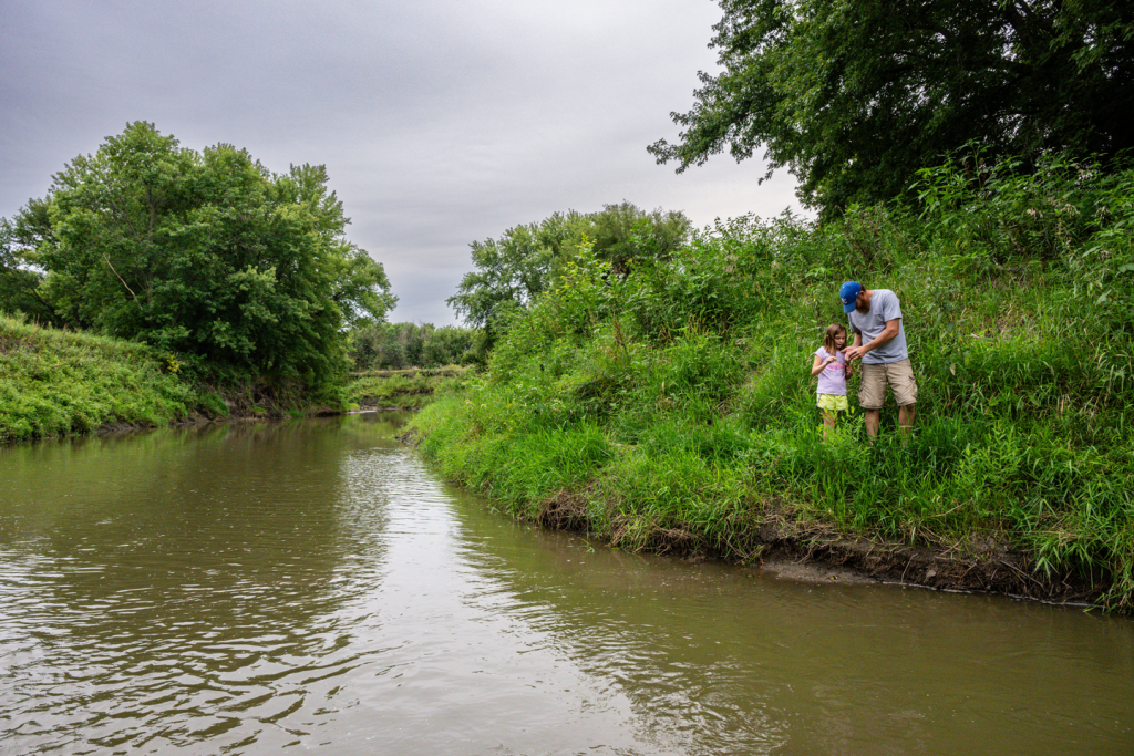 A father and child search for insects along the banks of a river during summertime.