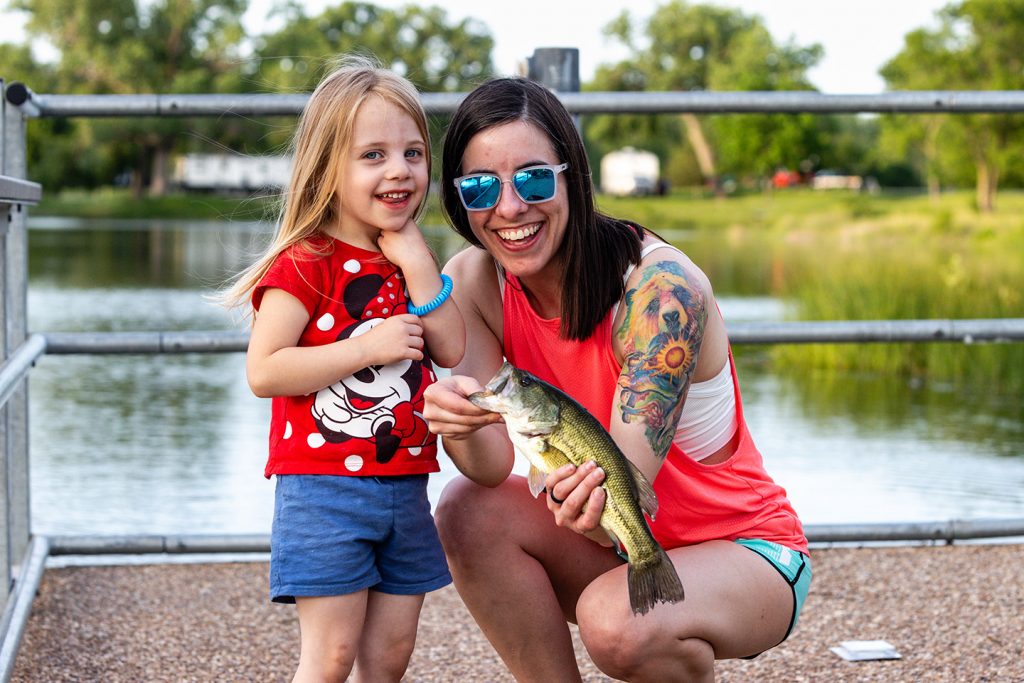 A woman and young girl smile and pose with a largemouth bass caught while fishing on a dock.