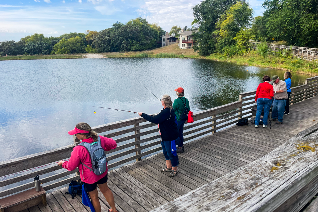 Women fish from a pier at a pond in a park.